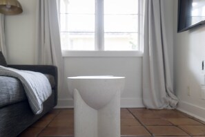 Una habitación moderna con una mesita de noche blanca, un sofá gris y una ventana con cortinas.