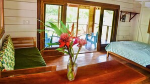 Une pièce avec un plancher en bois, un lit recouvert d’une couverture verte, un banc agrémenté d’un coussin à motifs et un vase rempli de fleurs.