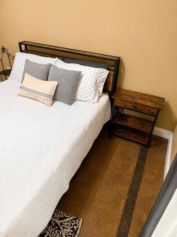 A bed with a white bedspread and multiple pillows, a wooden nightstand with drawers, and a patterned rug on a carpeted floor.