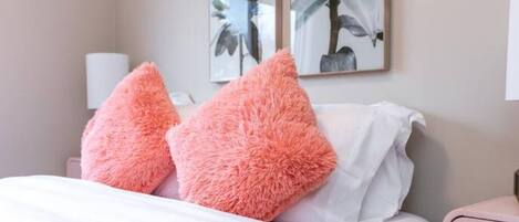 Ein Schlafzimmer mit einem Bett, zwei flauschigen rosa Kissen, weißer Bettwäsche und zwei Wandleuchten.