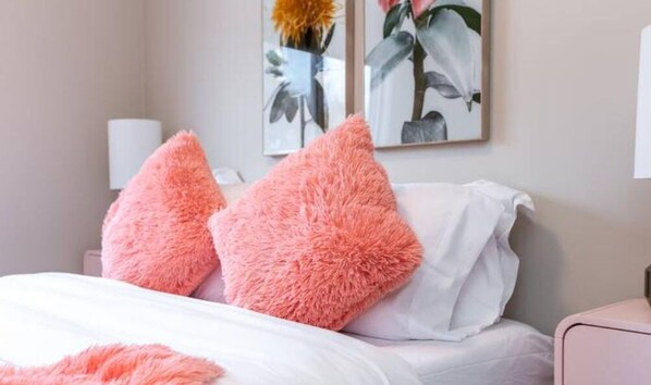 A bedroom with a bed, two pink fluffy pillows, white bedding, and two wall-mounted lamps.