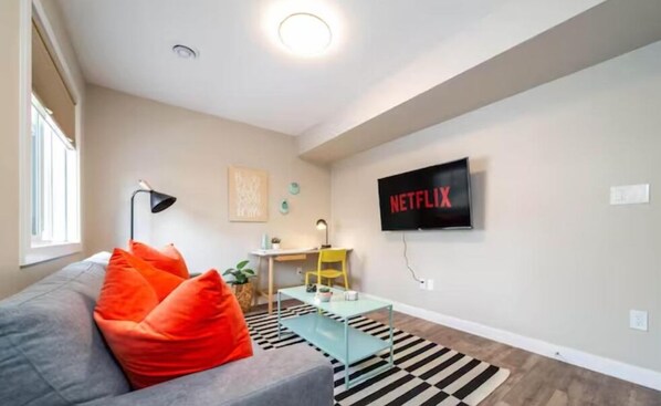 En moderne stue med en grå sofa, orange puder, et lille bord, en gul stol, en stribet måtte i sort og hvid og et vægmonteret fjernsyn med Netflix-logo.