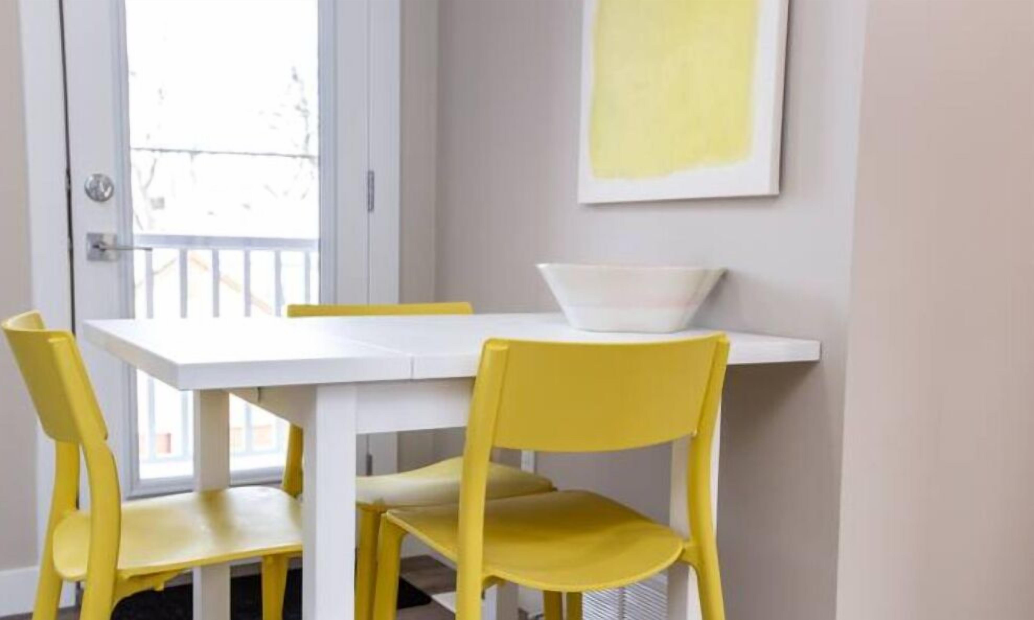 A room with a white table and yellow chairs, a white bowl on the table, and a yellow painting on the wall.