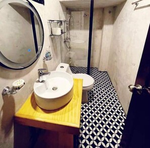 Une salle de bain avec un miroir rond, un lavabo doté d’un plan de toilette en bois, des toilettes et une douche. Le sol est recouvert de carreaux aux motifs géométriques noirs et blancs.