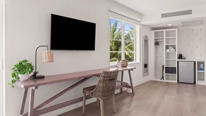 Een moderne kamer met een houten bureau, een rieten stoel, een aan de muur bevestigde televisie, een raam met uitzicht op palmbomen en een ingebouwde boekenkast met opbergruimte.