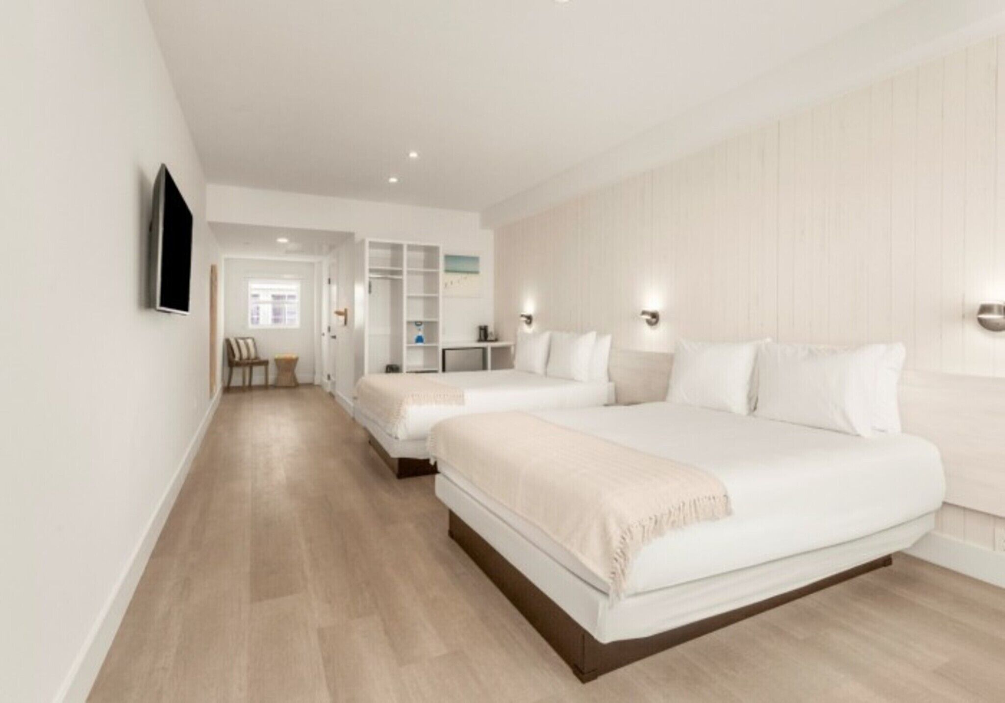 Habitación de hotel con dos camas, un televisor montado en la pared, piso de madera y pared blanca.