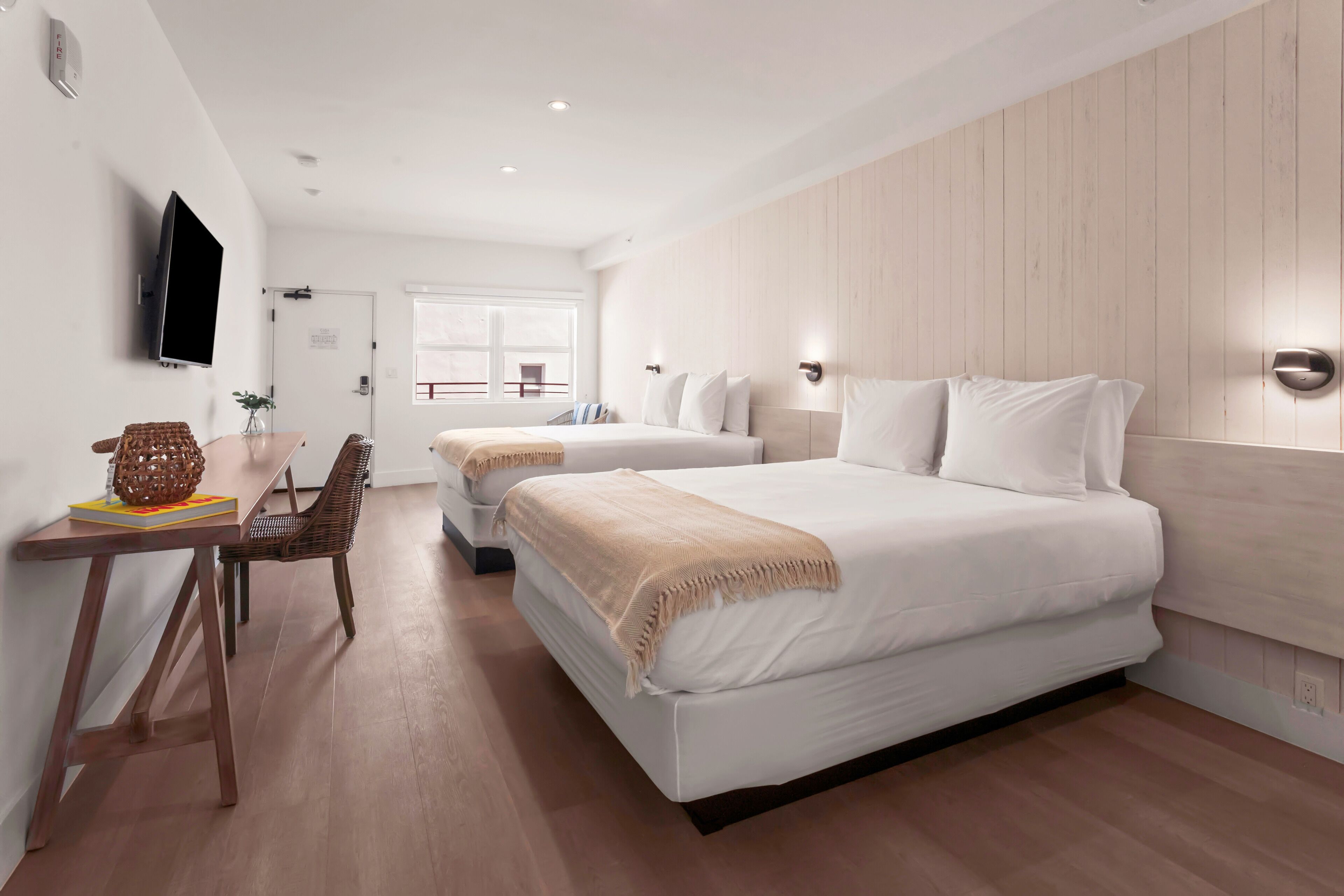 A modern hotel room with two beds, a wooden floor, a desk with a chair, and a TV mounted on the wall.