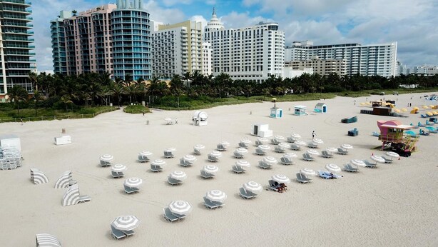 A beach with rows of white beach umbrellas and lounge chairs, a lifeguard tower, and high-rise buildings in the background. - Casa SOBE (Miami Beach)