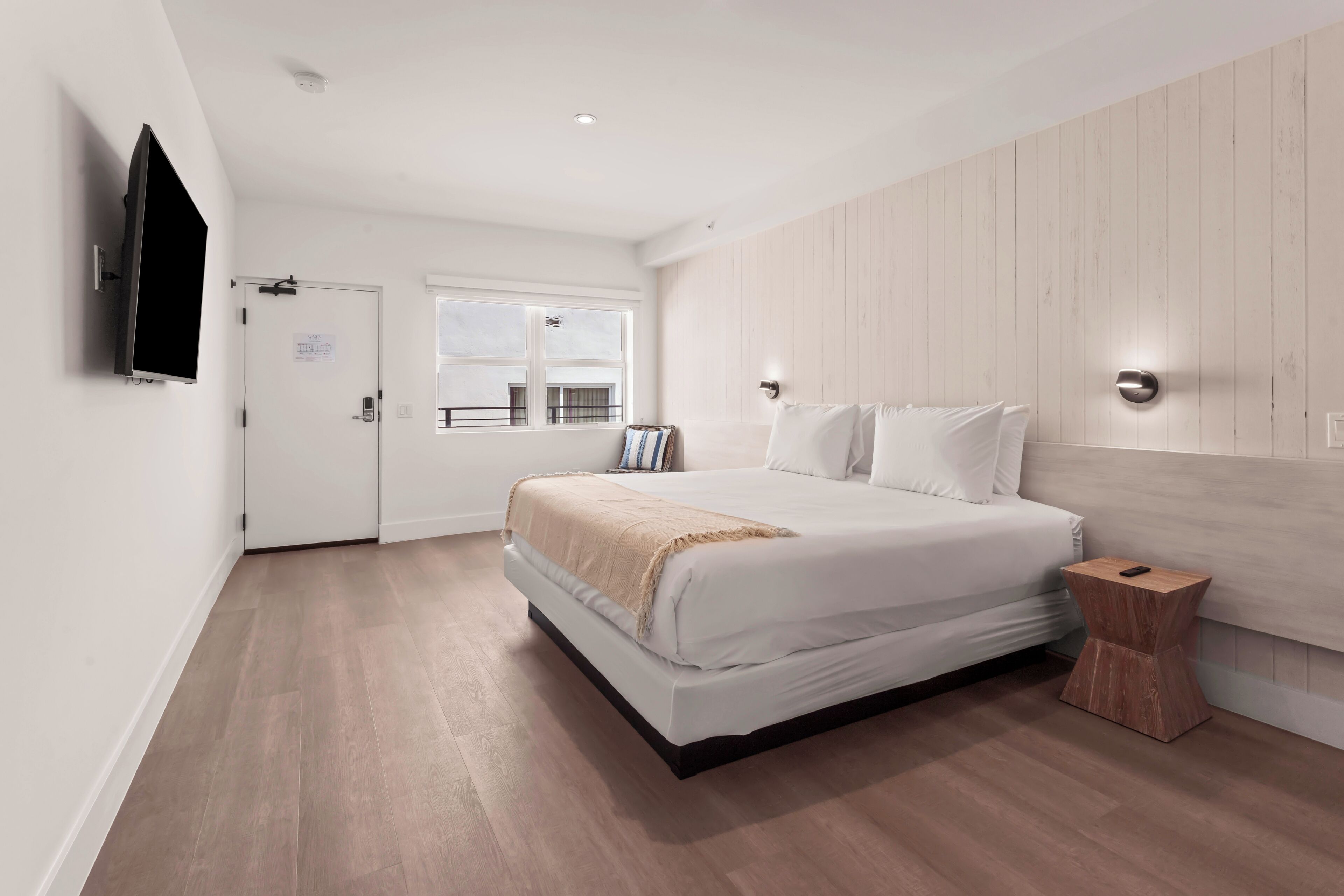 Une chambre moderne avec un grand lit, une table de chevet, un téléviseur fixé au mur et une fenêtre donnant sur l’extérieur.