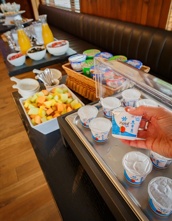 Una mesa de buffet con diversos alimentos y bebidas, incluyendo una bandeja con vasos de yogur y un bol de fruta.
