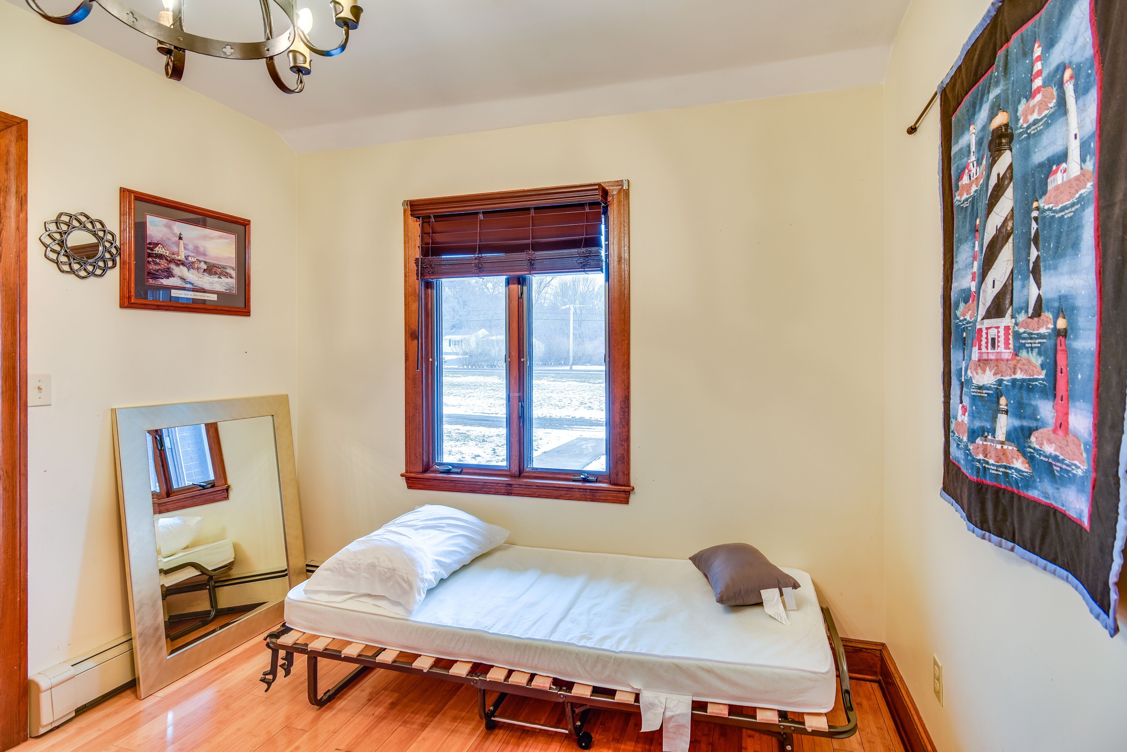 A small room with a single bed, a window with blinds, a mirror, and a wall with a framed picture.