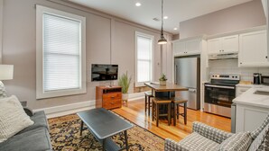 Un salon moderne avec un canapé gris, une table basse et un tapis à motifs. On y trouve également un coin repas avec une table et des chaises en bois. La cuisine est équipée de meubles blancs, d’un réfrigérateur en acier inoxydable et d’un four.