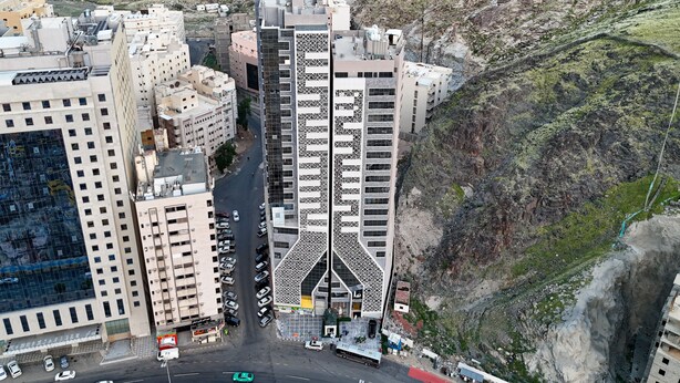 A high-rise building with a unique geometric facade, surrounded by other buildings and a steep hillside. - Lamar Ajyad First Hotel Tower B (Makkah)
