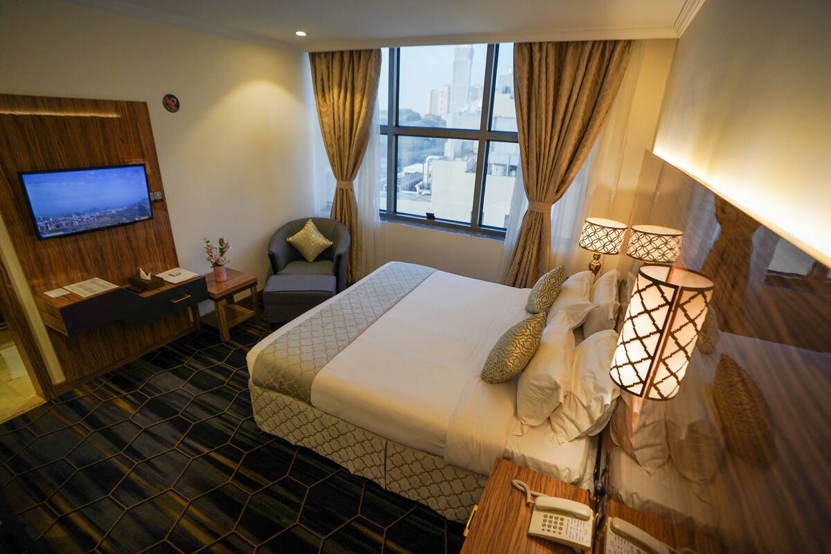 A hotel room with a bed, a desk with a TV, a chair, a window with curtains, and a lamp.