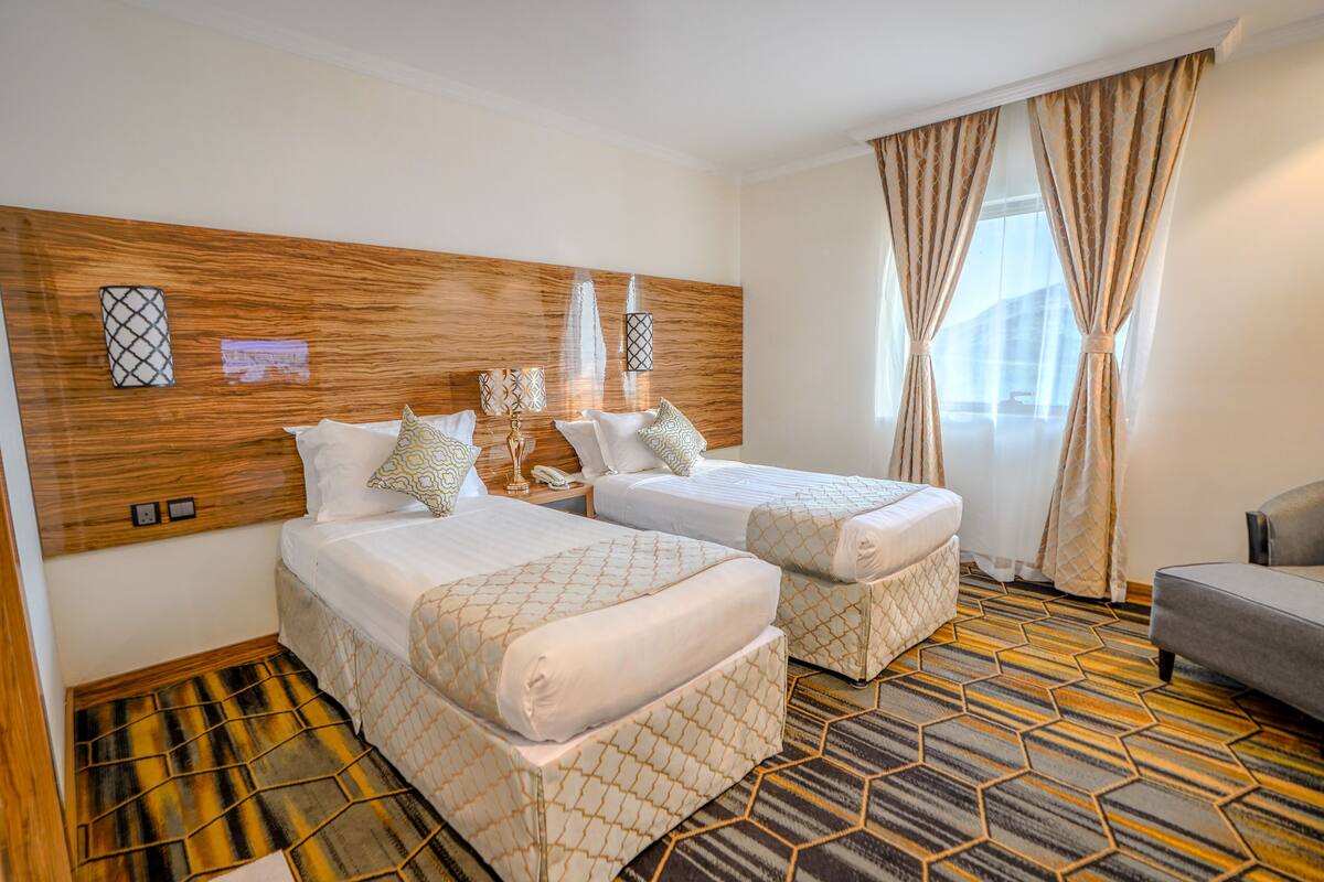 A hotel room with two beds, a wooden headboard, patterned carpet, and a grey armchair.
