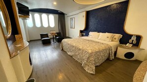Une chambre d’hôtel moderne avec un grand lit, un bureau et une chaise. Elle est dotée de fenêtres avec des rideaux, d’une petite table et d’un téléphone fixé au mur.