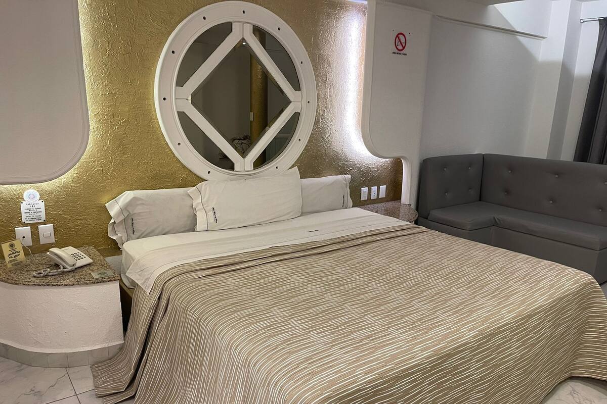 Une chambre d’hôtel moderne avec un grand lit, un miroir rond, un canapé gris et une petite table de chevet sur laquelle se trouvent un téléphone et un panneau.