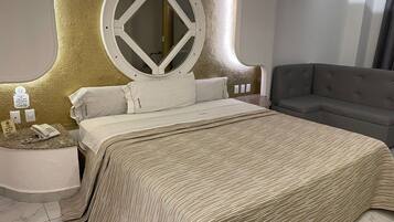 Una habitación de hotel moderna con una cama grande, un espejo redondo, un sofá gris y una mesita de noche pequeña con un teléfono y un cartel.