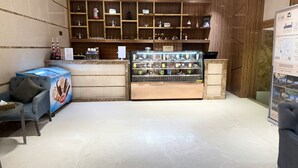 Interior de uma cafeteria com um balcão exibindo diversas bebidas, uma área de estar com um sofá azul e prateleiras de madeira com itens relacionados a café.