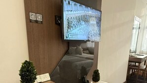 Quarto de hotel moderno com uma grande televisão de tela plana fixada num painel de madeira acima da cama. Há duas mesinhas de cabeceira com plantas decorativas.
