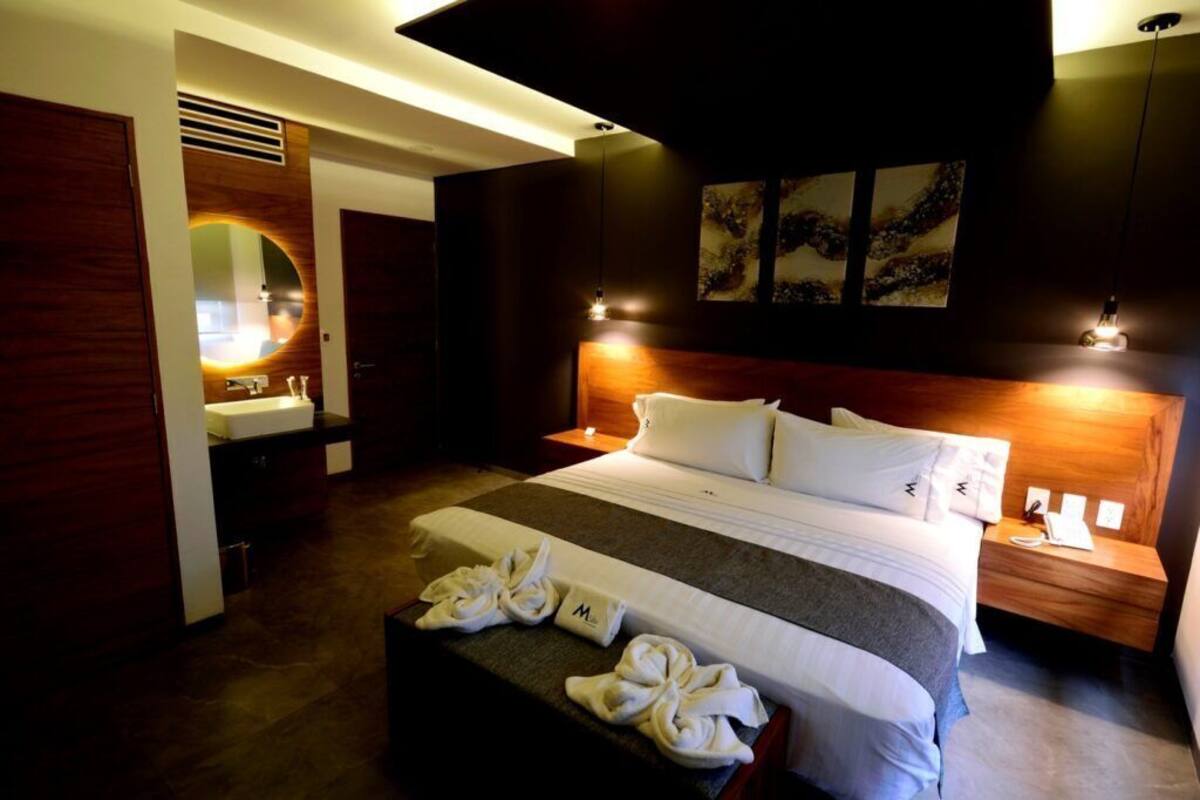 A modern hotel room with a large bed, a bedside table with a lamp, a mirror, and artwork on the wall.