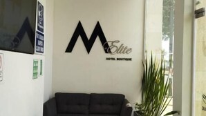 Een moderne hotellobby met een groot 'M Elite' bord, een zwarte bank, een klein rond tafeltje en een potplant.