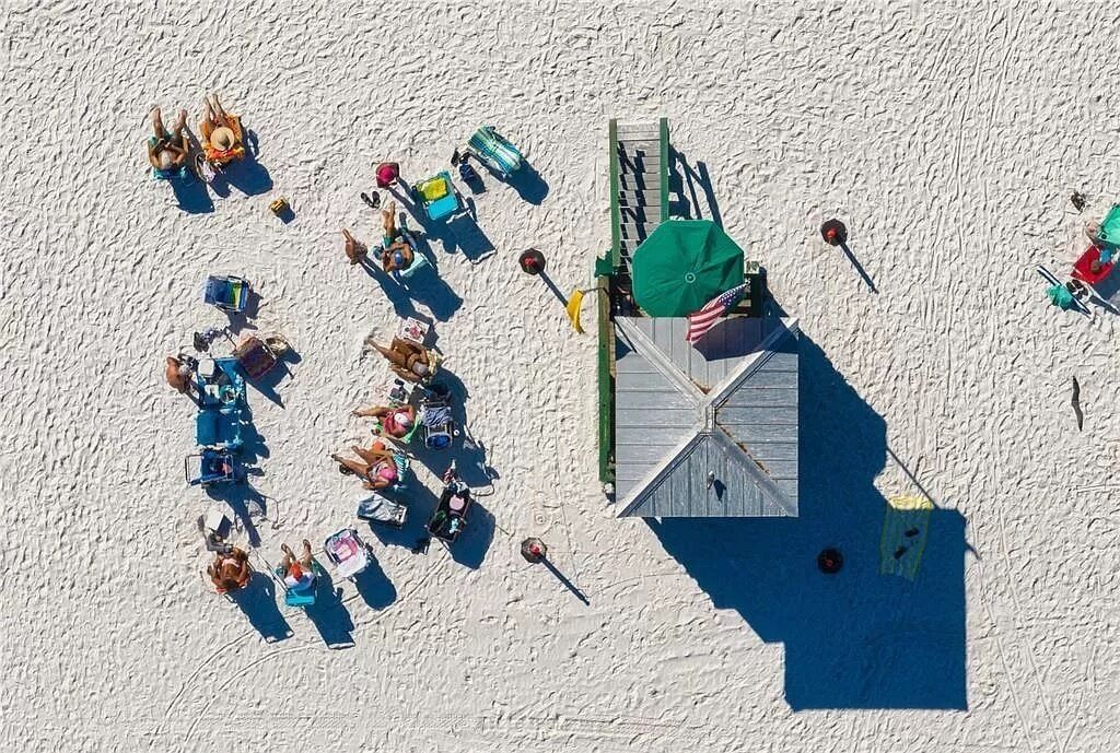 Vue aérienne d’une plage où l’on voit des gens se prélasser au soleil et nager, ainsi qu’une tour de surveillance et des chaises de plage.