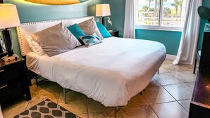 A neatly made bed with white linens, a teal-colored wall, a surfboard decoration, and a view of palm trees through a window. - Good Vibrations- Stay right ON the beach in Siesta Key! Sleeps 6 (Siesta Key)