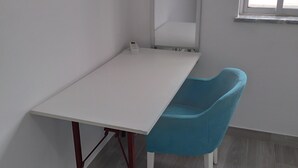 Una habitación con un escritorio blanco, una silla azul, un espejo grande y una ventana.