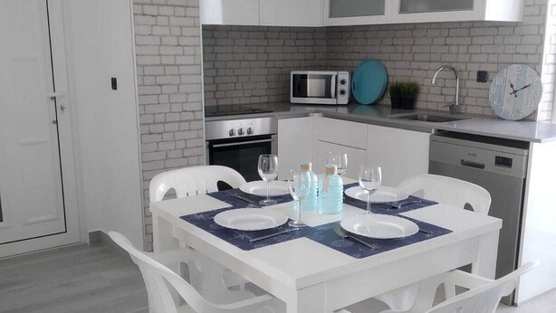 Una cocina moderna con armarios blancos, microondas, estufa y pileta. Hay una mesa de comedor con sillas y un reloj en la pared.