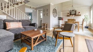 Uma sala de estar moderna com sofá cinza, mesa de centro de madeira e uma área de jantar com mesa e cadeiras de madeira. Uma escadaria é visível ao fundo.