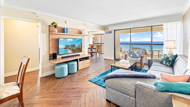 A spacious living room with a large TV, wooden flooring, a grey sofa, and a view of the ocean. - Oceanfront Condo W Free Valet Parking & Balcony (Honolulu)