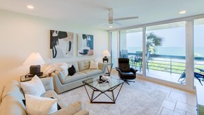 A modern living room with a sofa, coffee table, and armchair, large windows with a view of the ocean, and abstract art on the wall. - Premier Beach View 2 Bedroom Condo (Longboat Key)