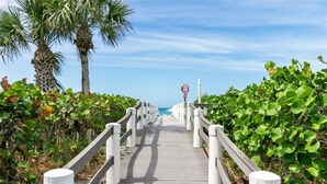 A wooden boardwalk with white railings, leading towards a body of water, surrounded by greenery and palm trees. - Premier Beach View 2 Bedroom Condo (Longboat Key)
