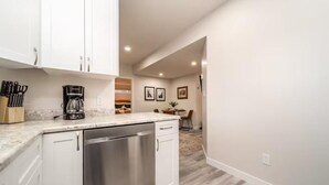 A modern kitchen with white cabinets, stainless steel dishwasher, and a coffee maker. There is a dining area with a table and chairs visible in the background. - NEW - Mountain Chic Escape - Whyte Ave - Netflix - Clean!
 (Edmonton)