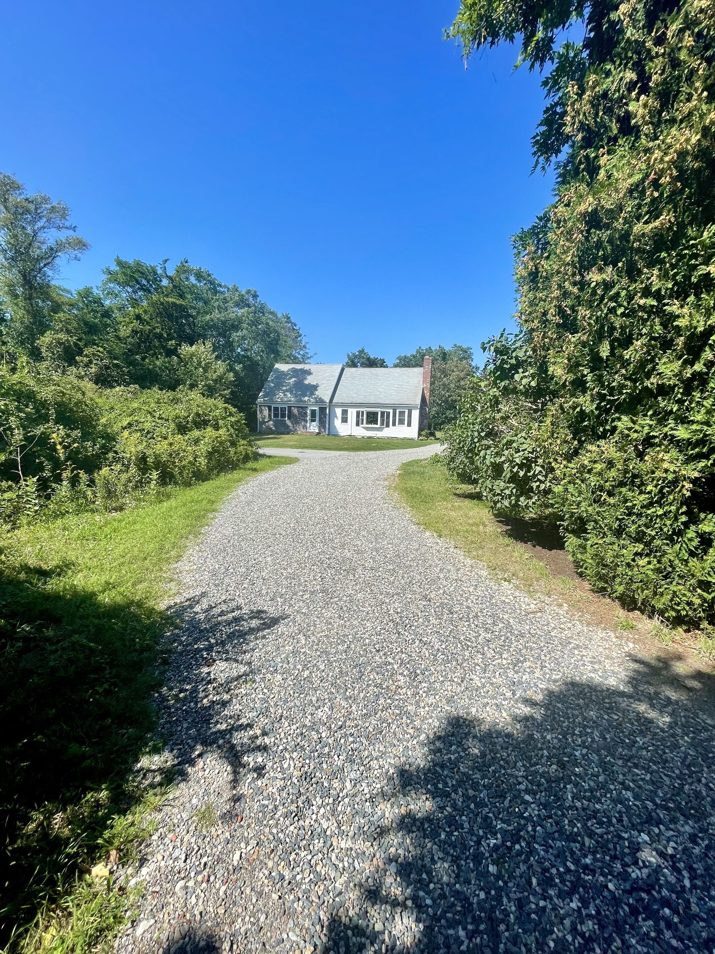 A gravel driveway leading to a white building with a sloped roof, surrounded by greenery.