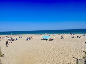Una extensa playa de arena con gente tomando el sol y paseando, bajo un cielo azul despejado.