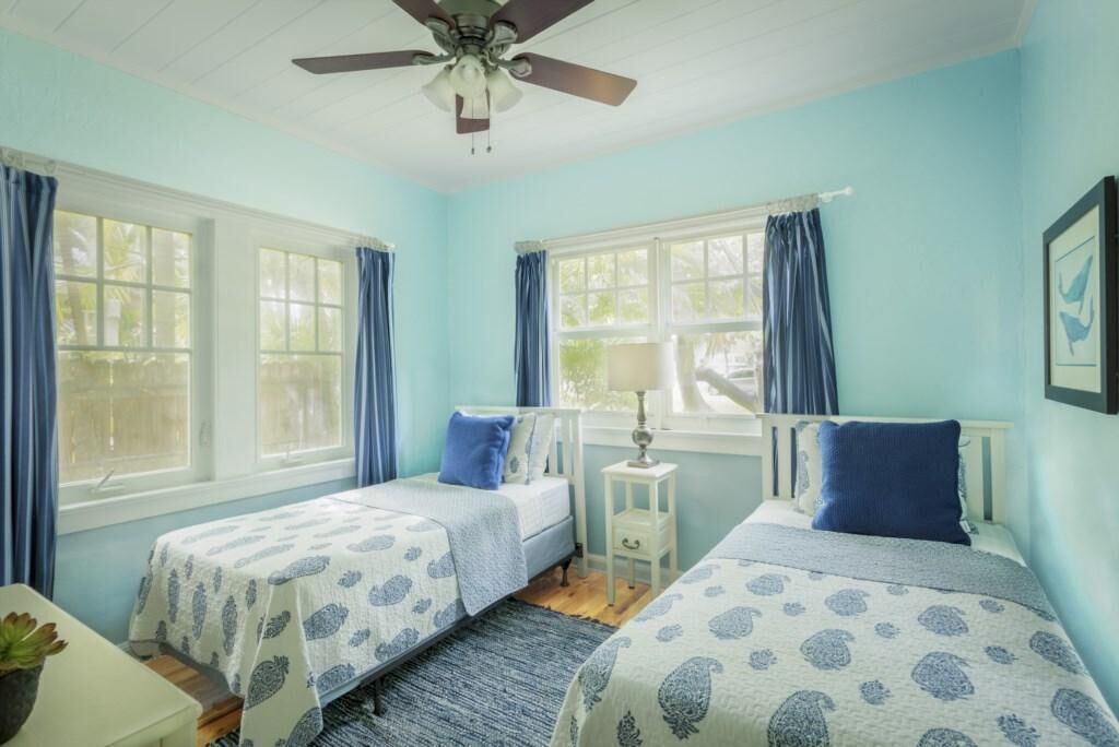 A room with two beds, a ceiling fan, and large windows with blue curtains.