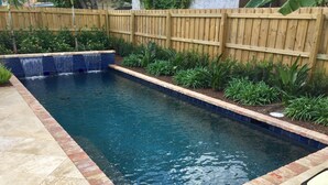 Una piscina rectangular con borde de azulejos, rodeada por un marco de ladrillo y una cerca de madera.