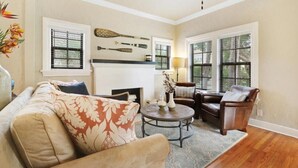 Un salón con sofá, sillón, mesa de centro, chimenea y grandes ventanales.