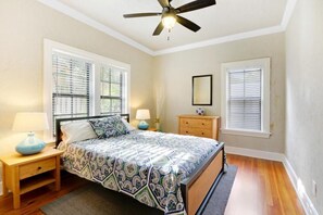A bedroom with a bed, two bedside tables, a ceiling fan, and two windows with blinds.