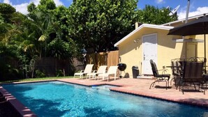 Une piscine rectangulaire bordée de briques, entourée de chaises longues et d’un espace patio aménagé avec une table et des chaises.