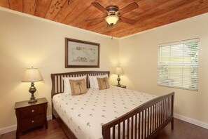 A bedroom with a wooden ceiling fan, a bed with a patterned bedspread, two bedside tables with lamps, a framed picture on the wall, and a window with blinds.