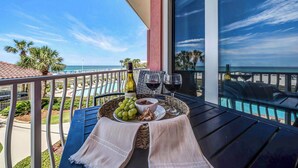 Uma varanda com uma mesa posta para dois, exibindo uma garrafa de vinho, uma tigela de chocolate e um prato de uvas. A varanda tem vista para uma piscina e palmeiras.