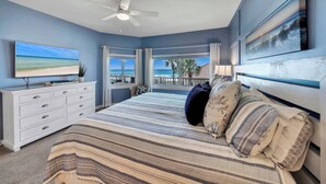 Quarto com uma cama grande, cômoda, ventilador de teto e vista para o oceano através de grandes janelas.