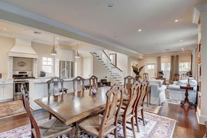 A spacious living area with a dining table, wooden chairs, a staircase, and a kitchen in the background.