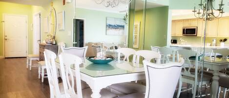 A dining area with a glass table and white chairs, a chandelier, and a kitchen visible through a glass partition.