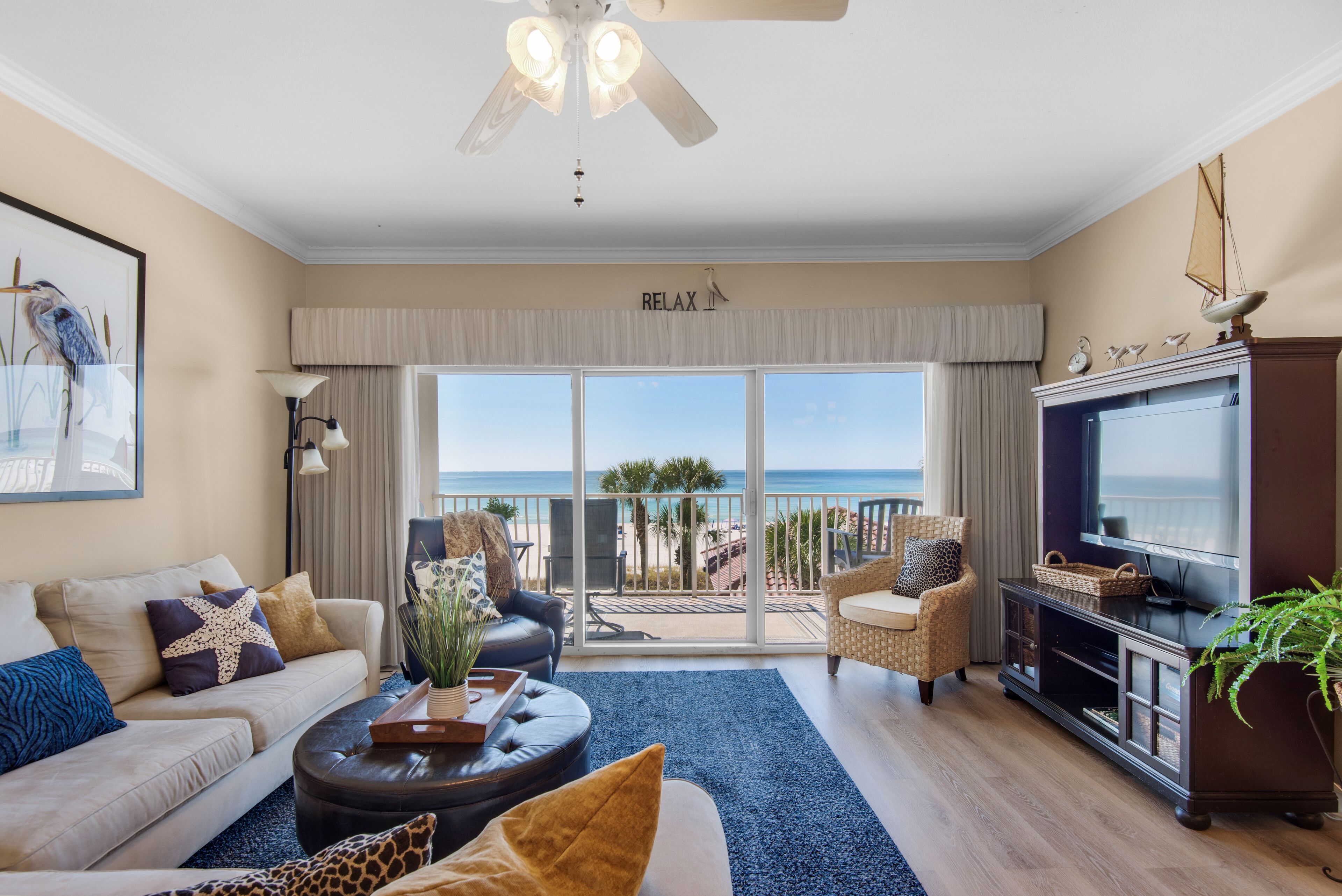 A spacious living room with a large TV, comfortable seating, a ceiling fan, and a balcony with a view of the beach. - Beachfront 2br/2ba condo on the 3rd floor with free beach chairs included! (Panama City Beach)