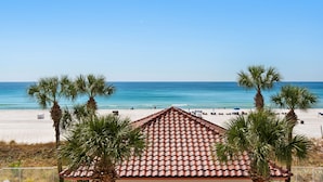 A beach view with a tiled roof building, palm trees, and a sandy beach. - Beachfront 2br/2ba condo on the 3rd floor with free beach chairs included! (Panama City Beach)