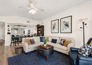 A living room with a sofa, coffee table, and chairs, a kitchen in the background, and two framed bird paintings on the wall.